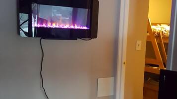 Habitación con televisor de pared mostrando un fuego, sistema de sonido y cama al fondo.