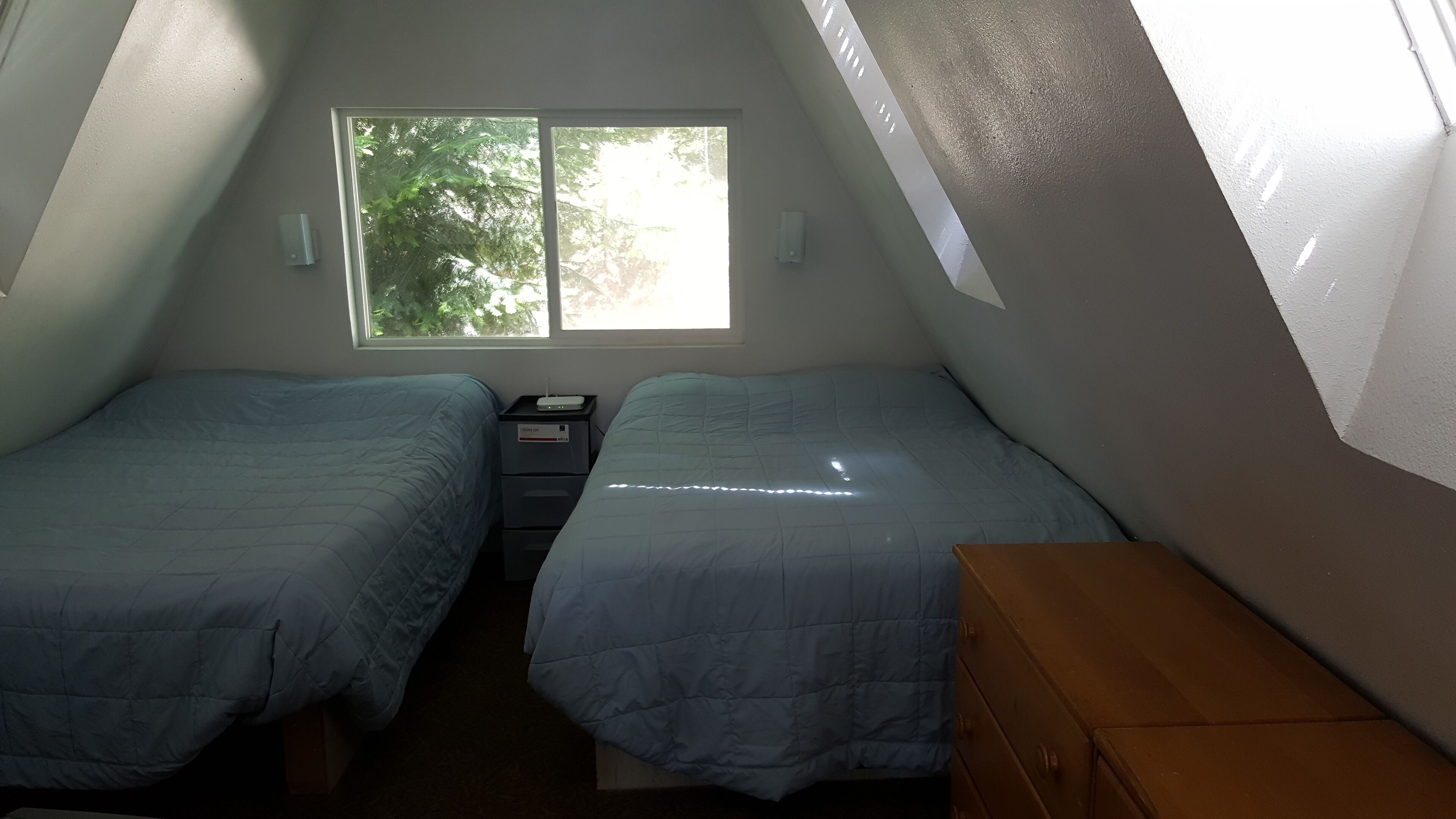 A small, triangular attic bedroom with two beds, a nightstand, a dresser, and a window with a view of trees.
