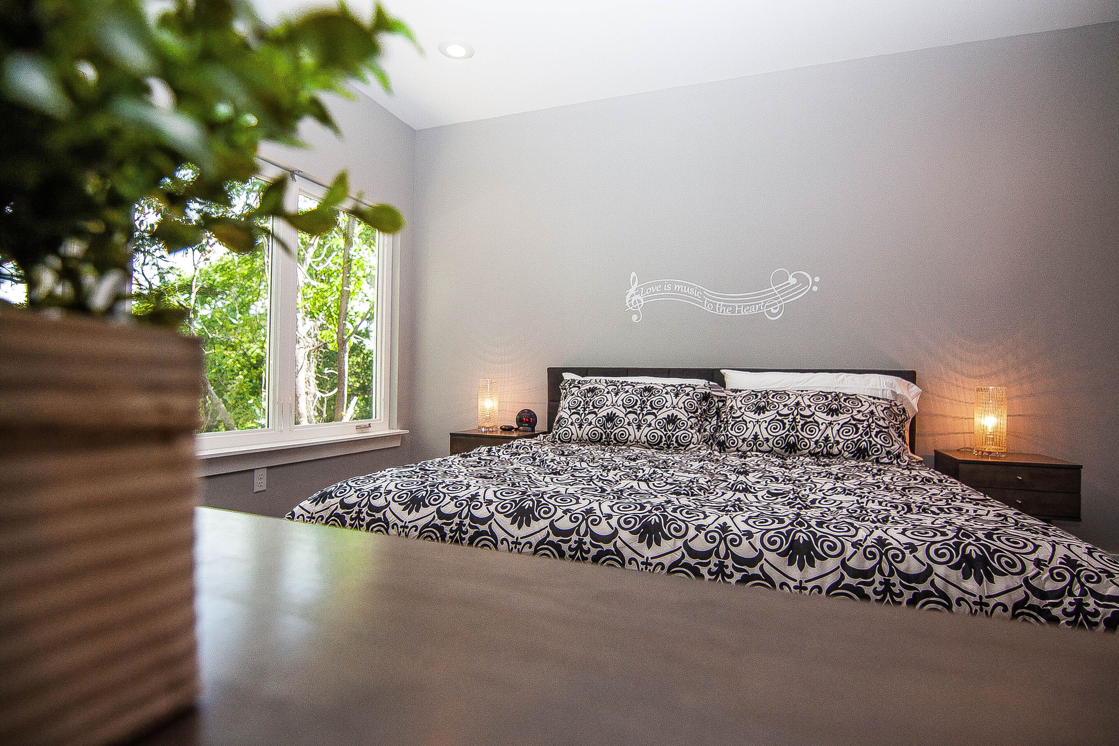 A bedroom with a patterned bedspread, a wall decoration, and a window with a view of trees.