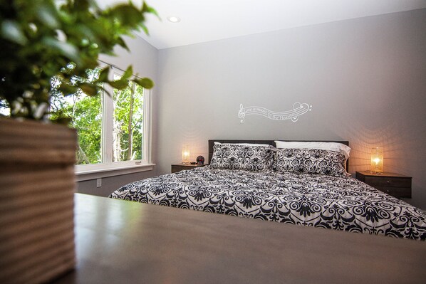 A bedroom with a large bed, two bedside tables, a wall decoration, and a window with a view of trees.