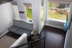 A modern living room with a grey sofa, a small wooden table with a plant, and a large window offering a view of a garden.
