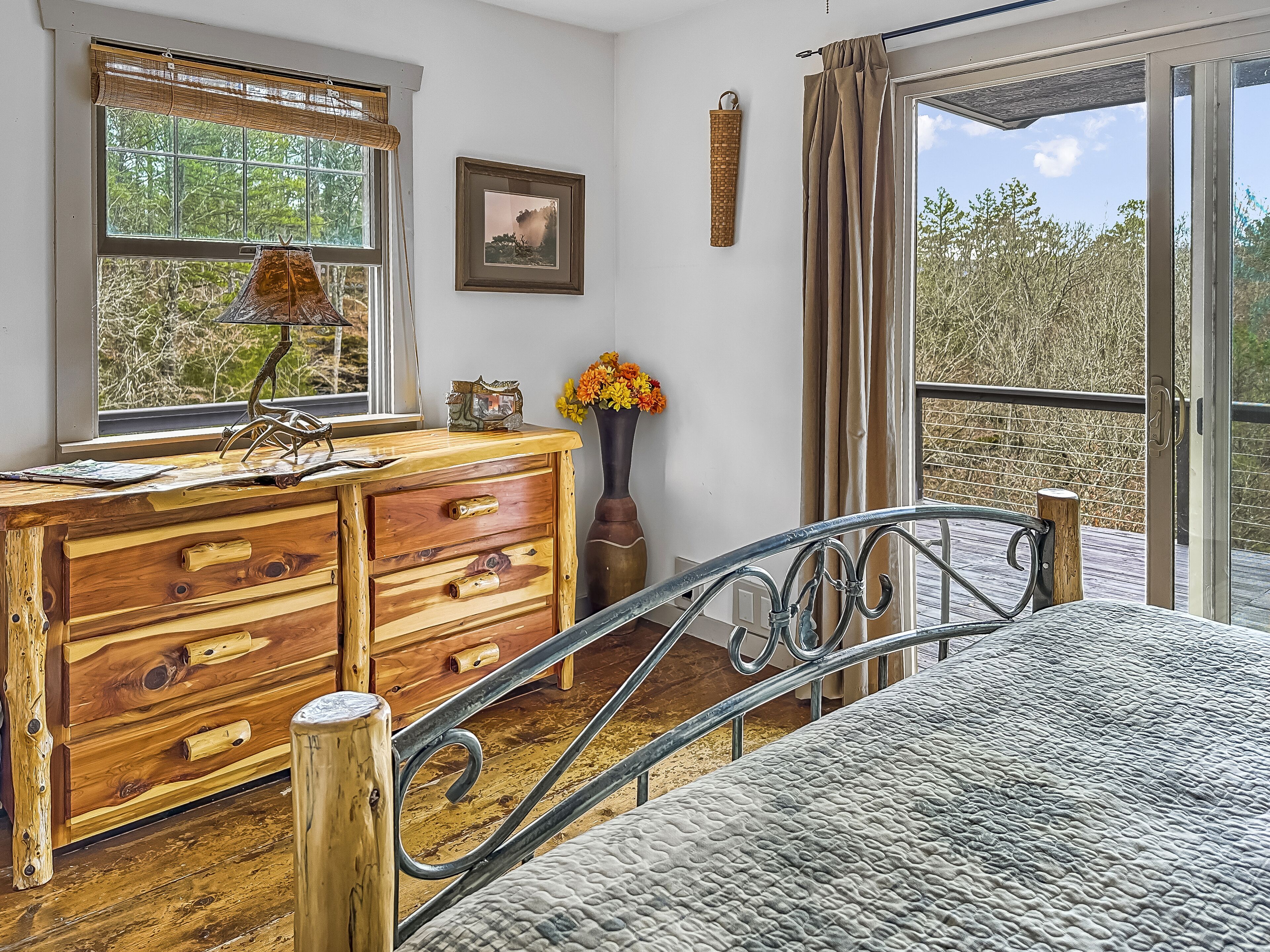 A bedroom with a wooden dresser, a bed with metal railings, a window with a view of trees, and a picture on the wall.