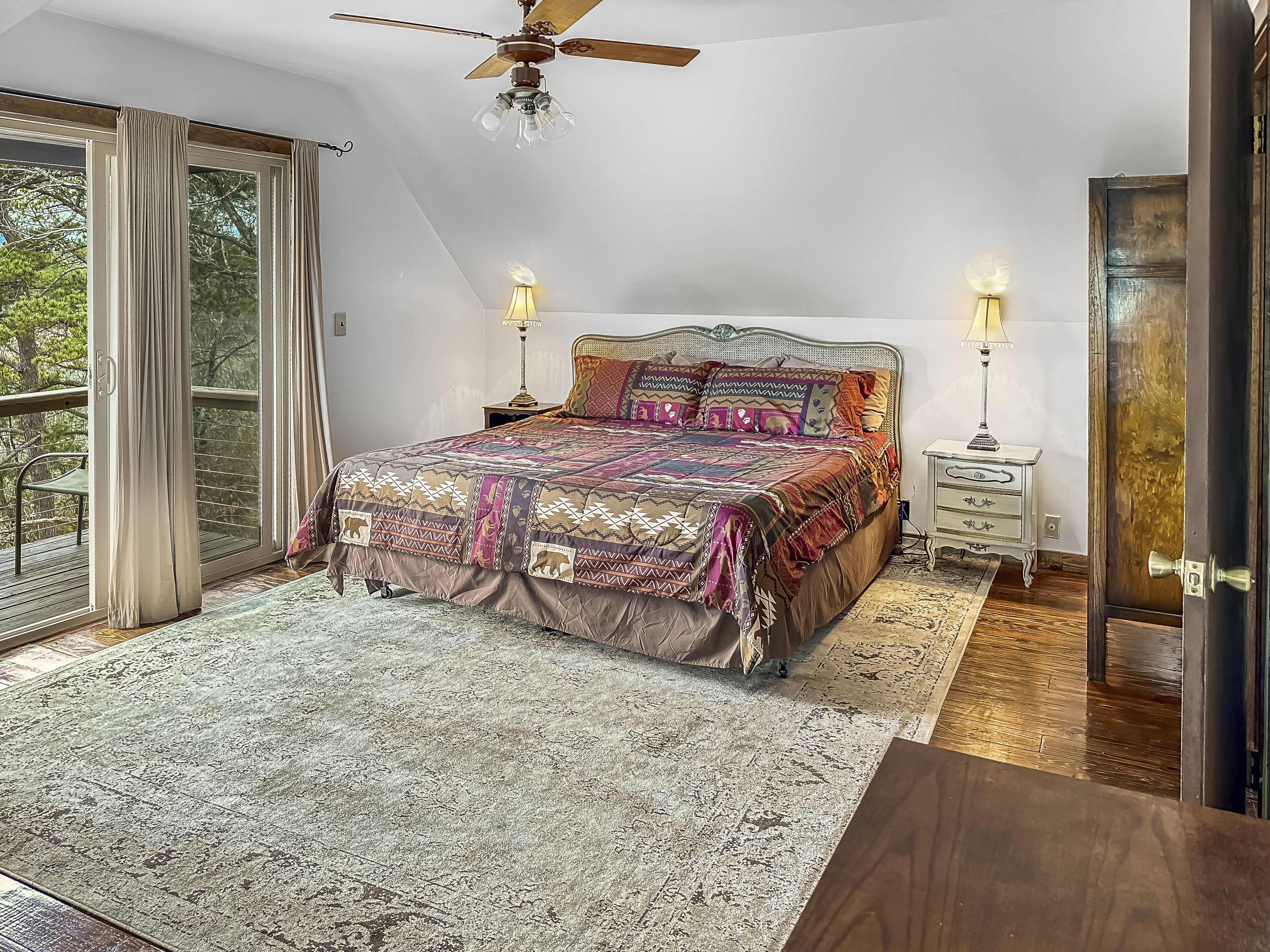 Une chambre avec un grand lit, deux lampes de chevet, un ventilateur de plafond et une vue sur l’extérieur, accessible par une porte-fenêtre coulissante.