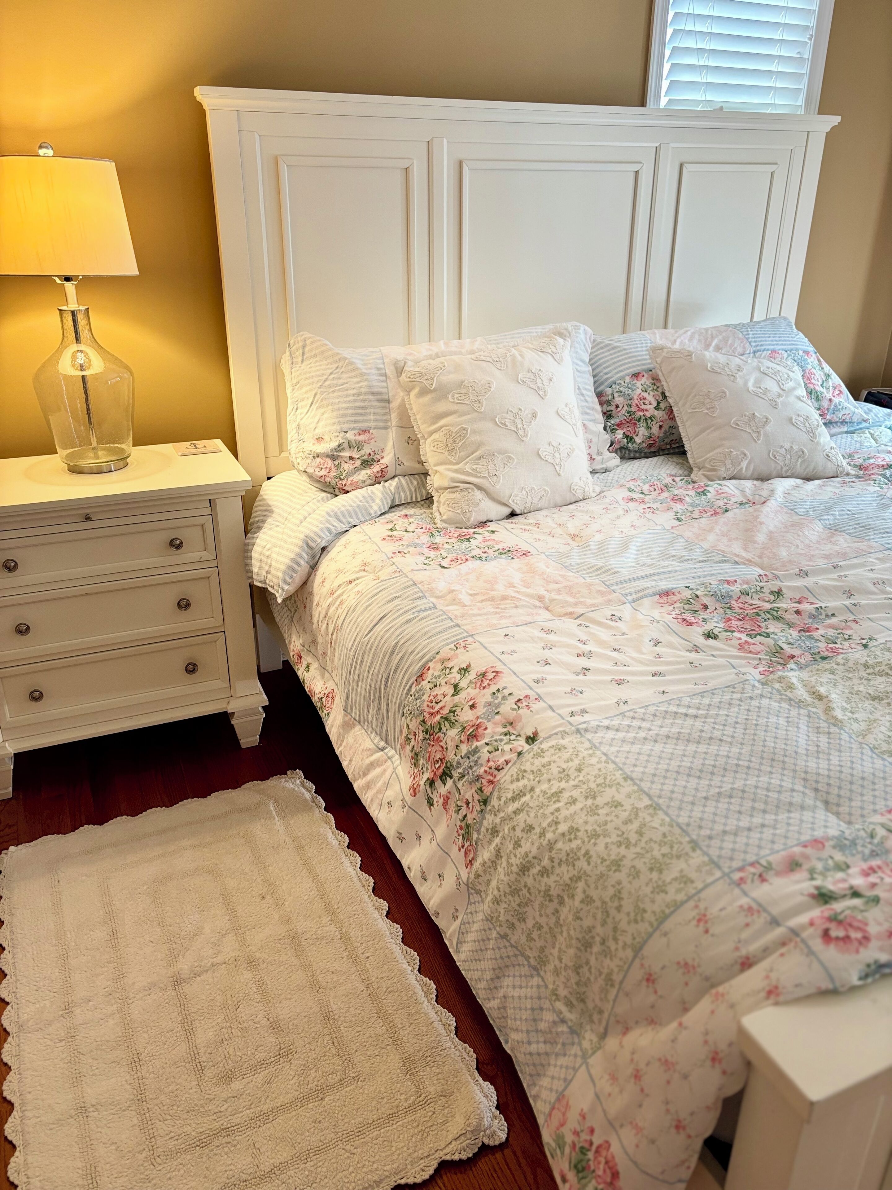 A bedroom with a white bed, floral bedding, a bedside lamp, and a white nightstand.