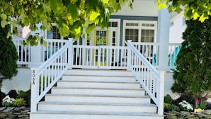 Eine weiße Treppe führt zu einer Veranda mit weißer Brüstung, umgeben von Grünpflanzen und einer Steinsäule.