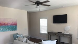 A living room with a ceiling fan, a flat-screen TV, a painting, a sofa, a desk, and a patterned rug. - Newer home 2 blocks from downtown Carolina Beach (Carolina Beach)