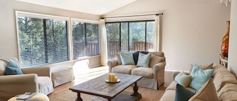 Ett rymligt vardagsrum med en hög takfläkt, stora fönster med persienner, en beige soffgrupp, ett soffbord i trä och en dekorativ vas.