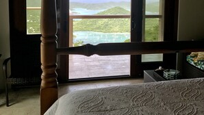 A bedroom with a large window offering a view of a coastal landscape, a bed with a patterned bedspread, and a wooden chair. - HEAVENLY VIEWS and PRIVATE POOL!! (Coral Bay)