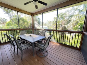 A covered outdoor patio with a table and chairs, surrounded by a wooden deck and metal railing, overlooking a lush garden. - The Osceola House at Indian Pass (Port St. Joe)