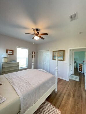 A bedroom with a bed, a ceiling fan, a window, a door, and a hallway leading to another room. - The Osceola House at Indian Pass (Port St. Joe)