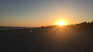A beach at sunset with the sun low on the horizon, casting a warm glow over the sand and water.