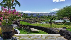 A tropical garden with a stone pathway, lounge chairs, and a view of a river and mountains. - Paradise Awaits! Gorgeous Kolea Condo with Great Views (Waikoloa)