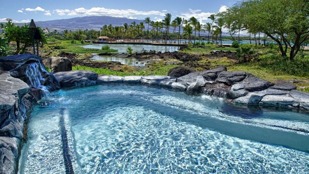 A hot spring with clear water, surrounded by rocks and palm trees, with a view of mountains and a pool in the background. - Paradise Awaits! Gorgeous Kolea Condo with Great Views (Waikoloa)