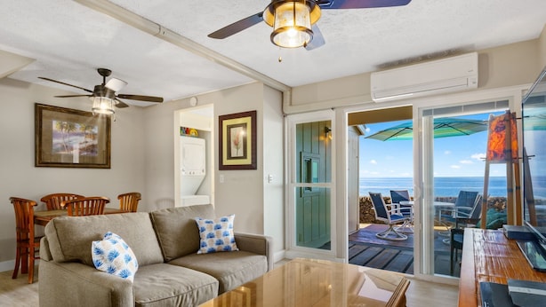 A living room with a sofa, coffee table, and ceiling fan. There is a door leading to a balcony with a view of the ocean. - Direct Oceanfront Condo on Private Beach Resort *Central AC * (Kailua-Kona)