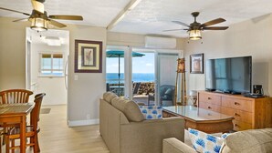 A living room with a sofa, TV, wooden dining table, chairs, ceiling fans, and a view of the ocean through a sliding glass door. - Direct Oceanfront Condo on Private Beach Resort *Central AC * (Kailua-Kona)