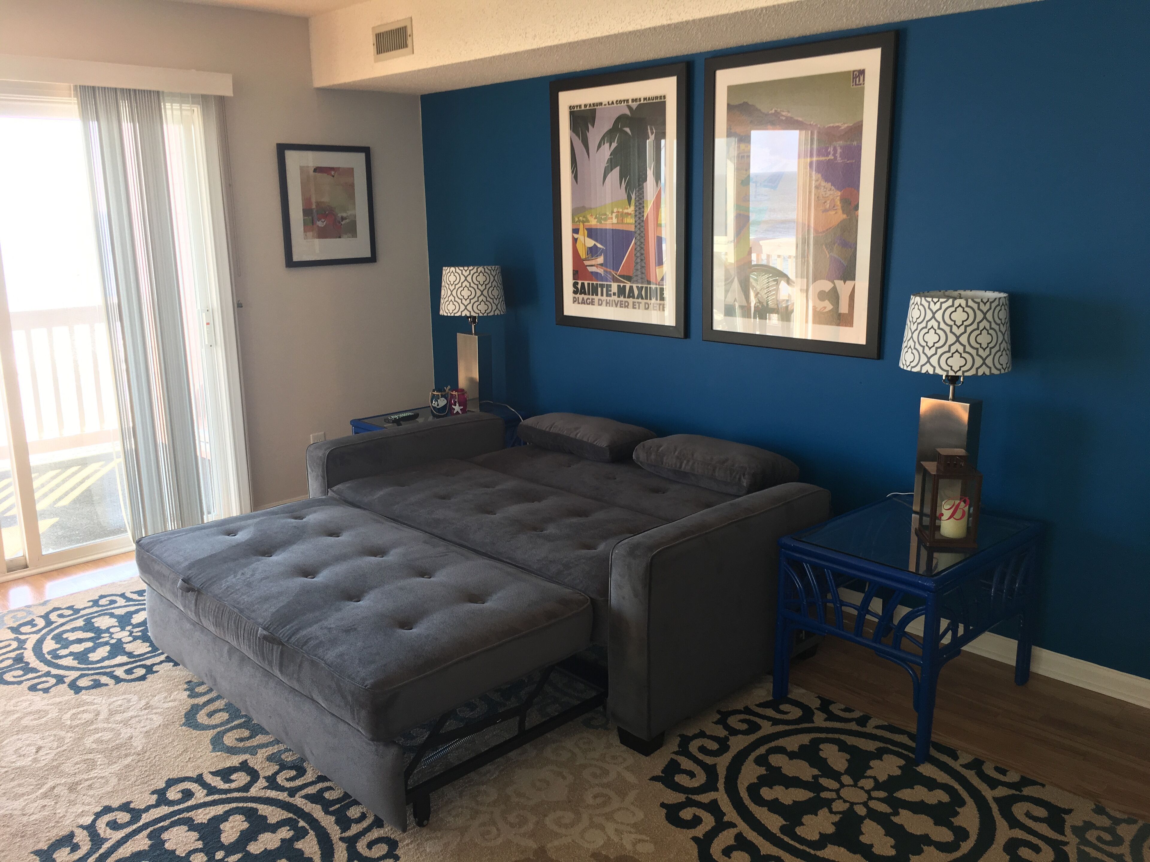 A bedroom with a grey sofa bed, a blue accent wall, two framed pictures, a patterned rug, and a small table with a lamp.