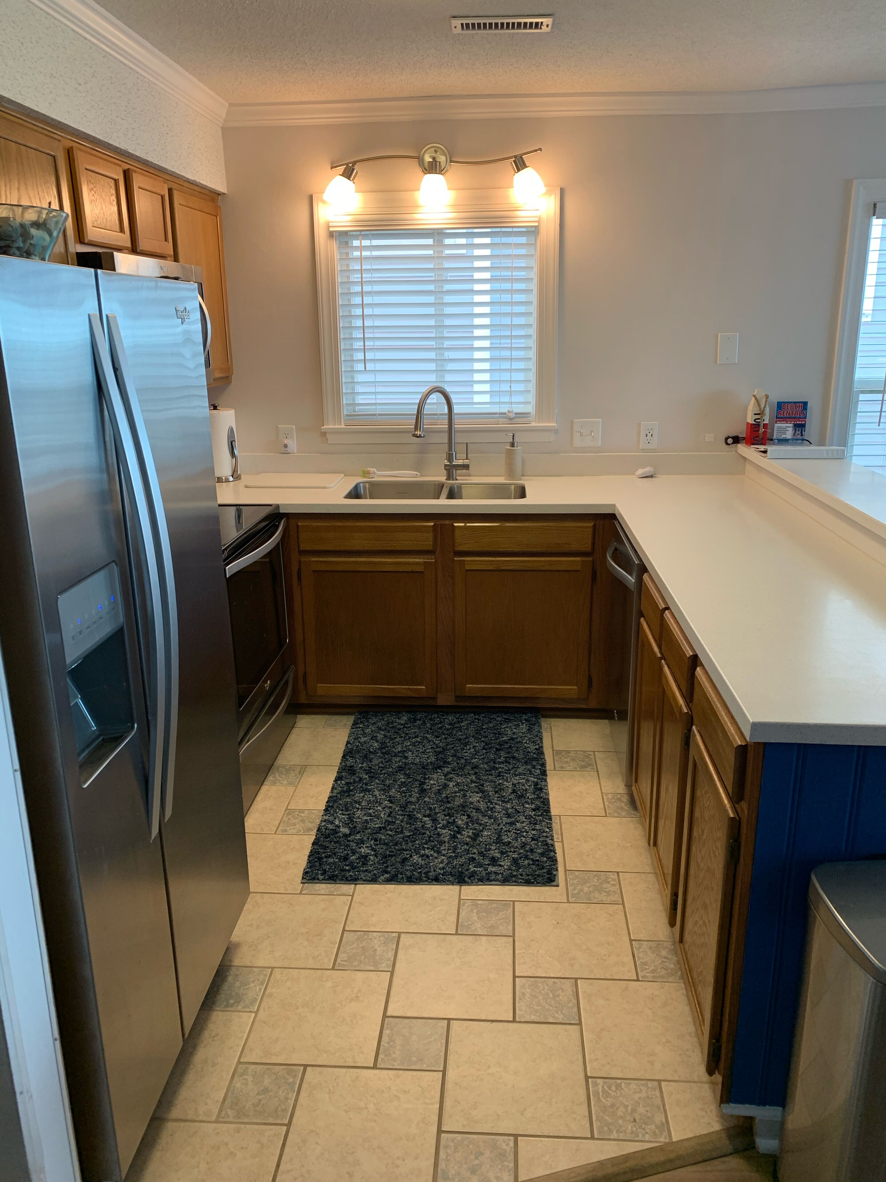Una cocina moderna con un refrigerador de acero inoxidable, un lavamanos doble, armarios de madera, una ventana con persianas y una alfombra azul oscuro.