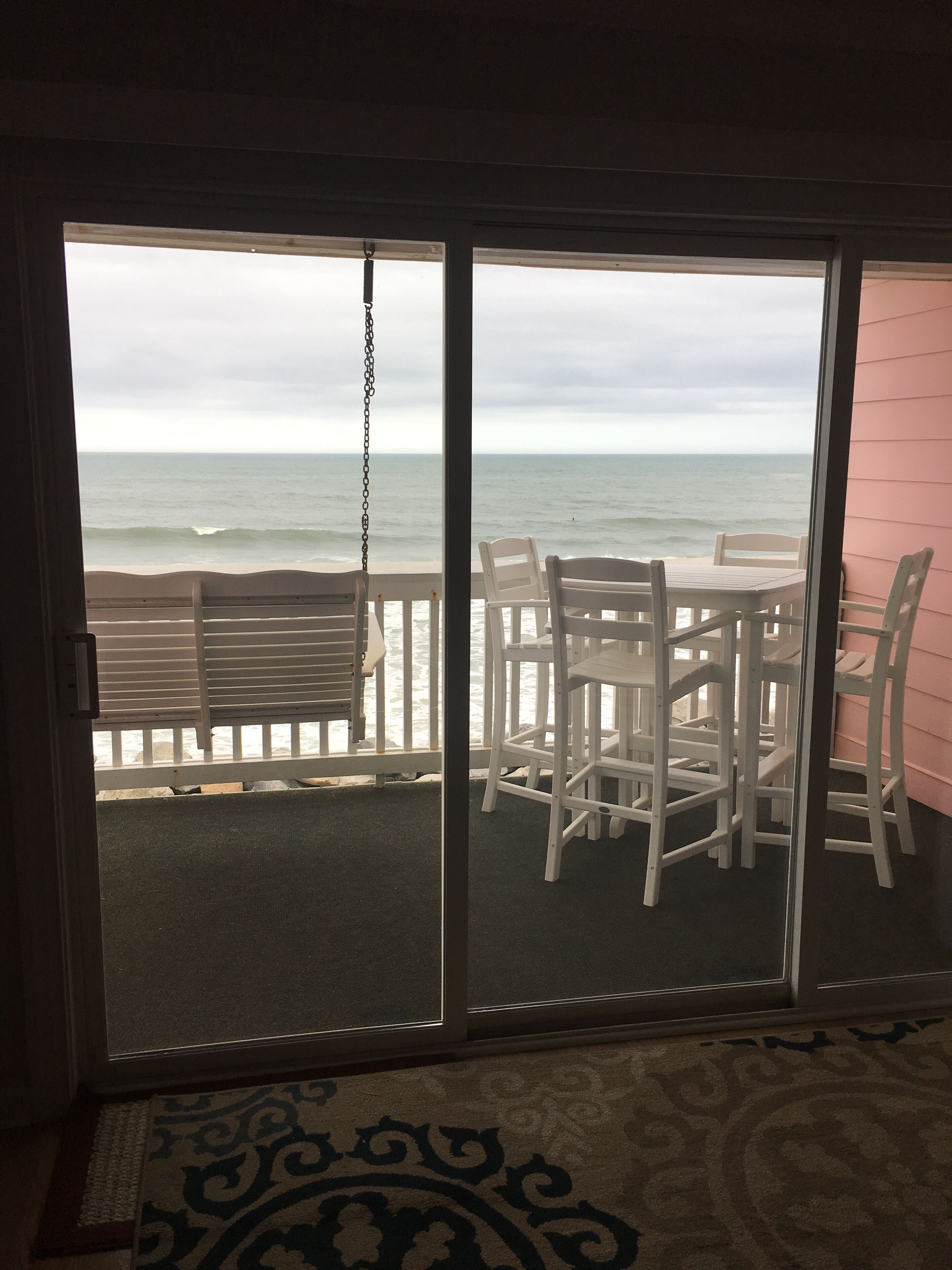 A room with a view of the ocean through a sliding glass door. There is a white table and chairs set up outside.