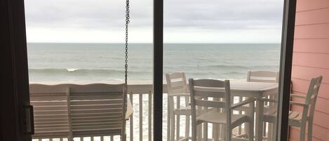 A room with a view of the ocean through a sliding glass door. There is a white table and chairs set up outside.