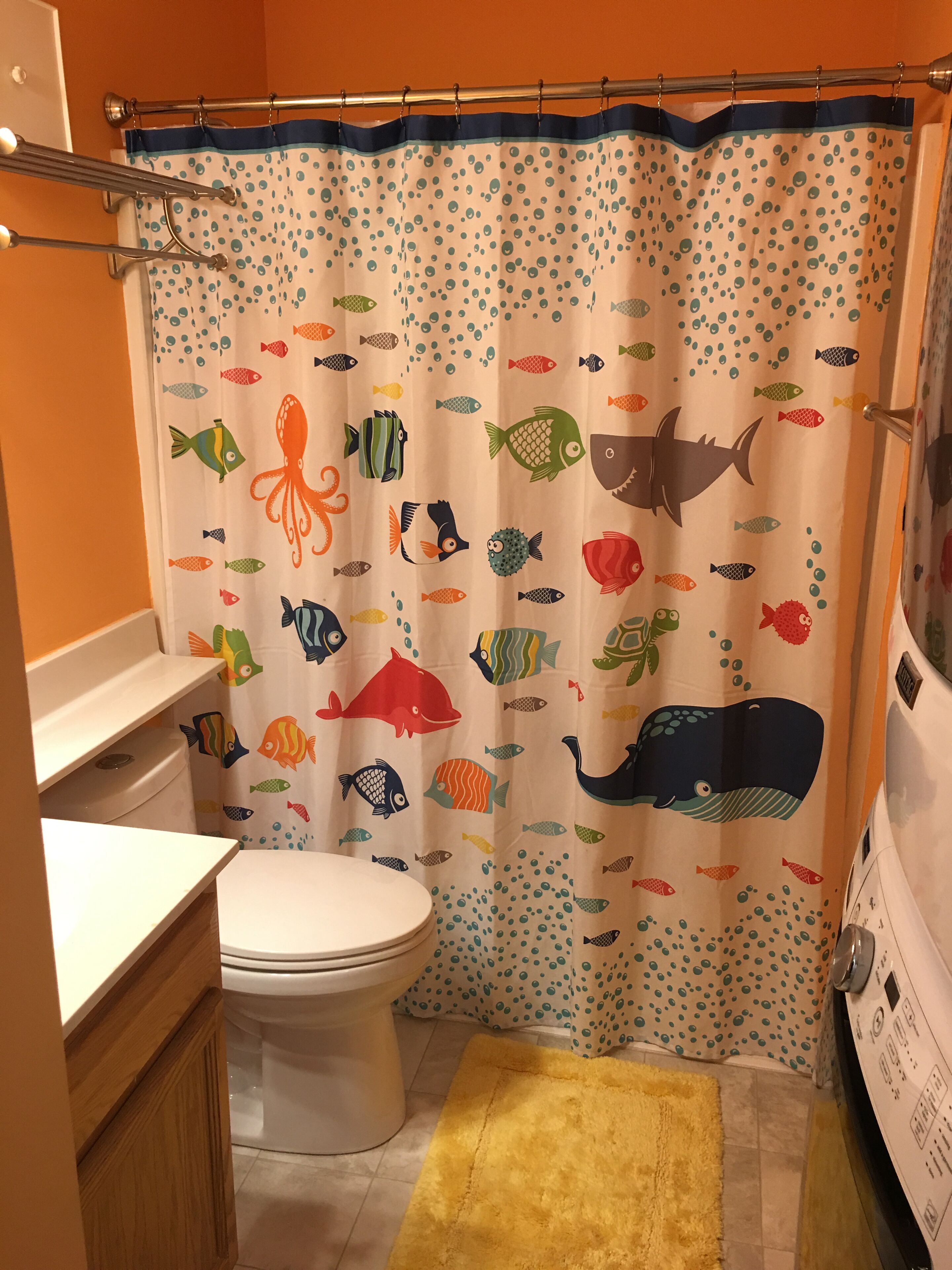 A bathroom with a toilet, a yellow rug, and a shower curtain with fish and whale patterns.