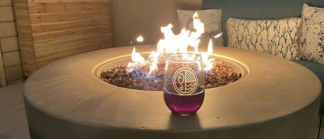 A round outdoor fire pit with a glass container in the center, surrounded by a wooden planter and a patterned sofa.