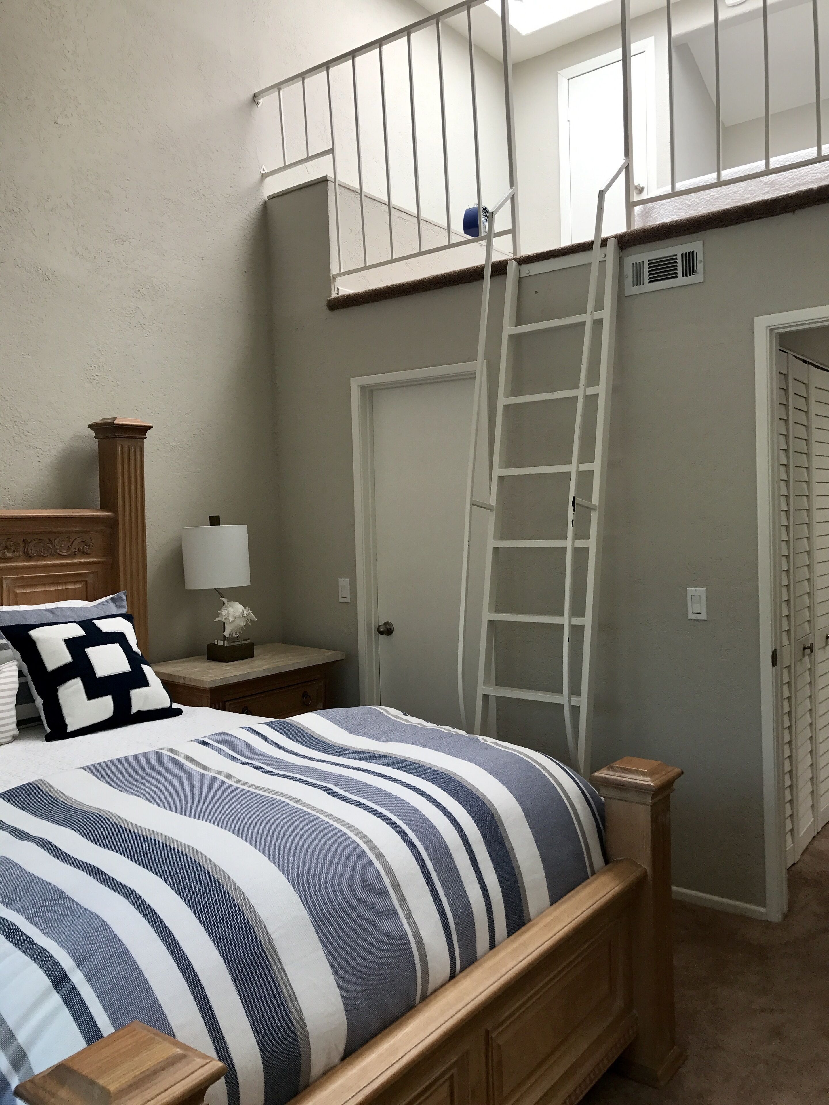 Quarto com cama, mesa de cabeceira, abajur, cômoda, escada e porta.