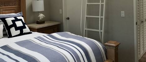 Quarto com cama, mesa de cabeceira, abajur, cômoda, escada e porta.