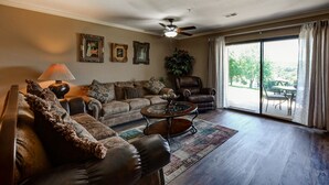 A living room with a sofa, armchair, coffee table, and a ceiling fan. There are two framed pictures on the wall and a potted plant.