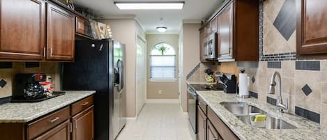 A kitchen with wooden cabinets, granite countertops, a refrigerator, microwave, and a window with blinds.
