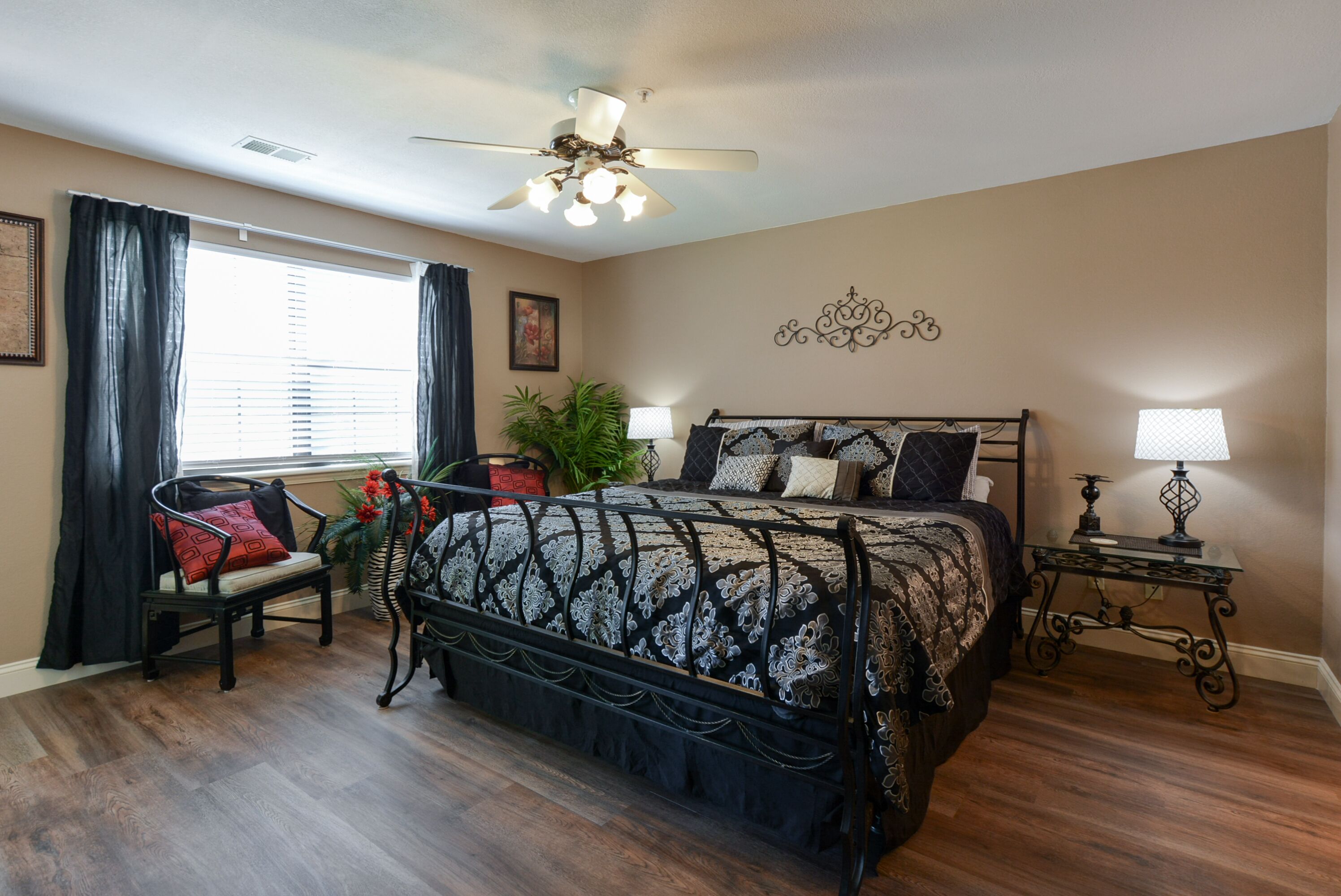 A bedroom with a large bed, a chair, a nightstand, a ceiling fan, and a window with curtains.