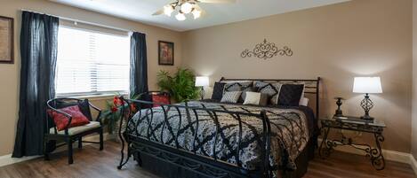 A bedroom with a large bed, a chair, a nightstand, a ceiling fan, and a window with curtains.