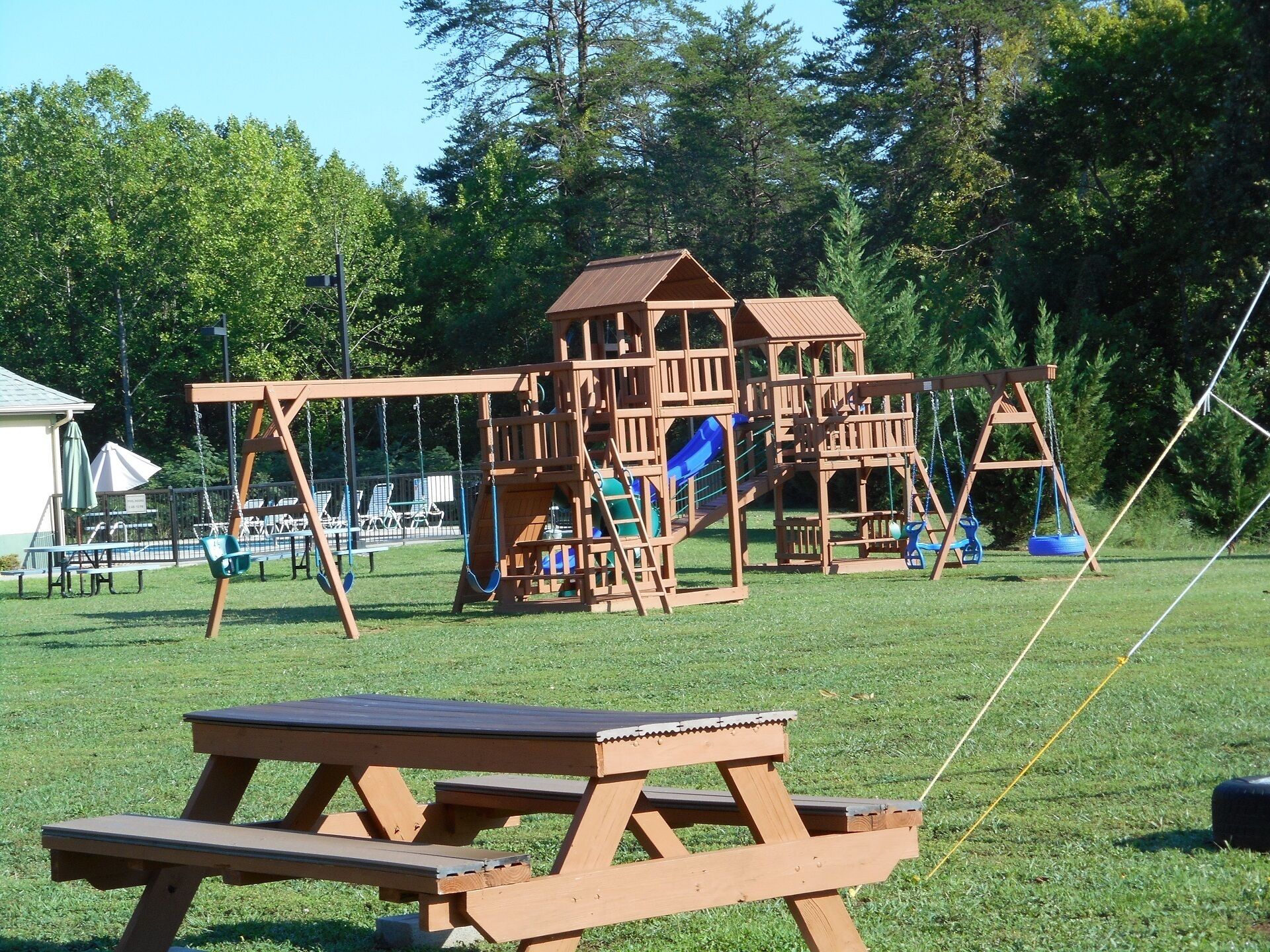 A wooden playground with slides and swings, a picnic table, and a fenced area with lounge chairs.