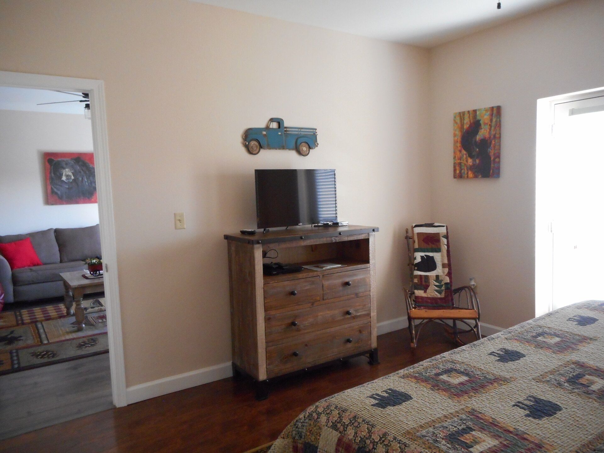 Une chambre à coucher avec un lit, une commode en bois, un téléviseur posé dessus, une chaise et une porte.