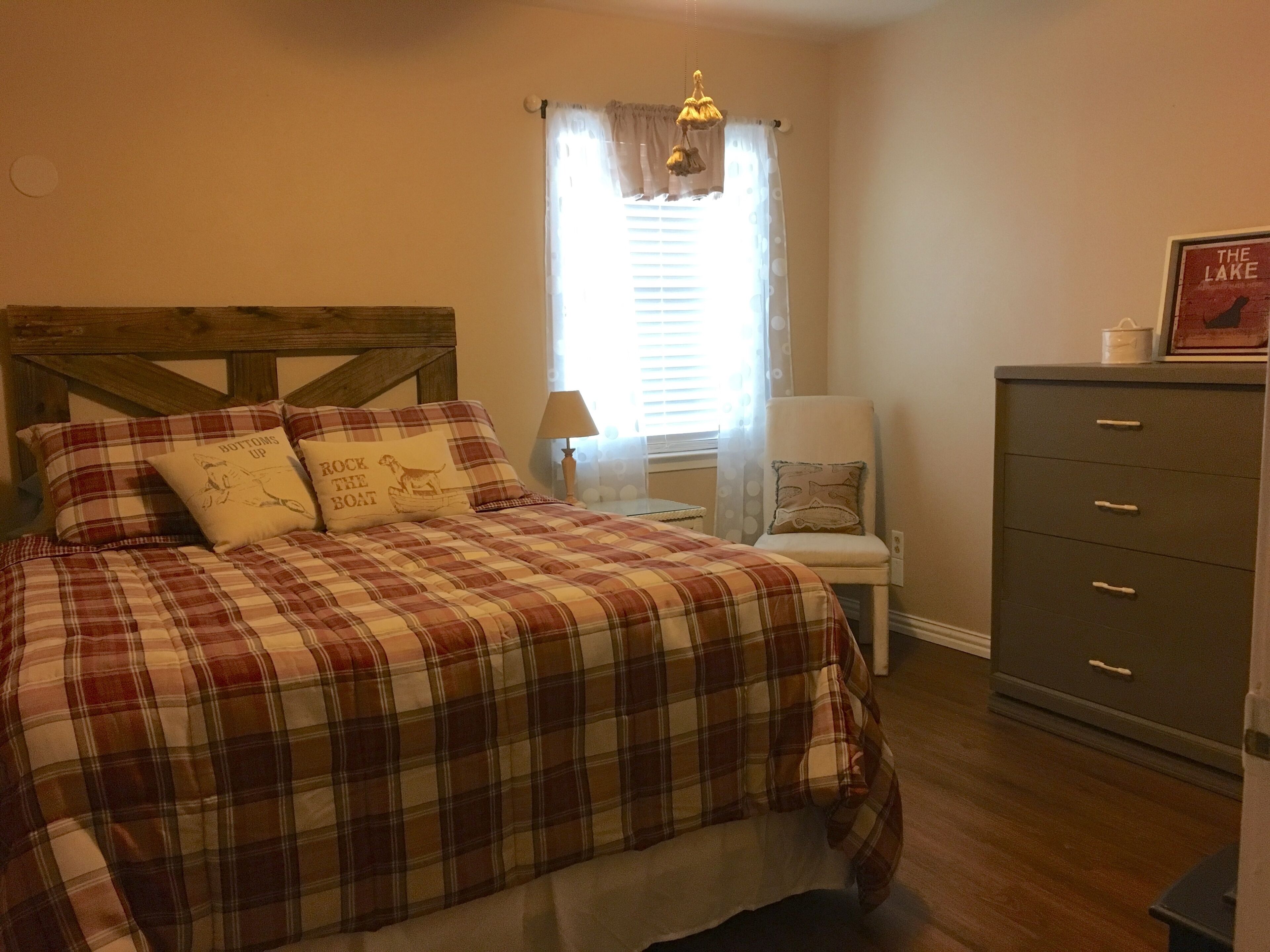 A bedroom with a plaid bedspread, a wooden headboard, a chair, a nightstand, a lamp, a dresser, and a window with curtains.
