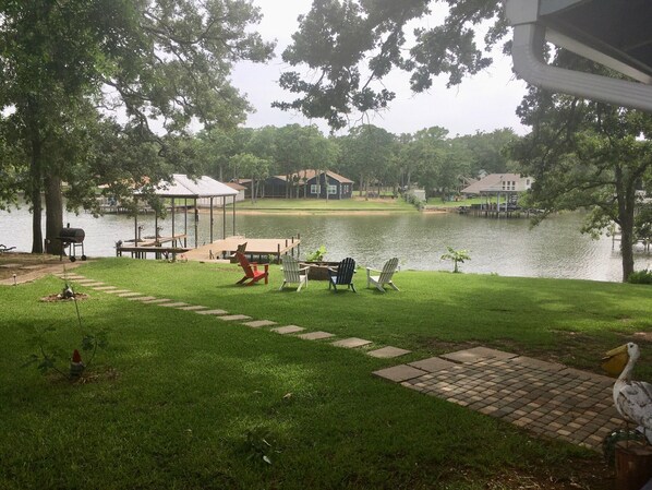 Un patio junto al lago con una parrilla, sillas y un muelle que se extiende sobre el agua.