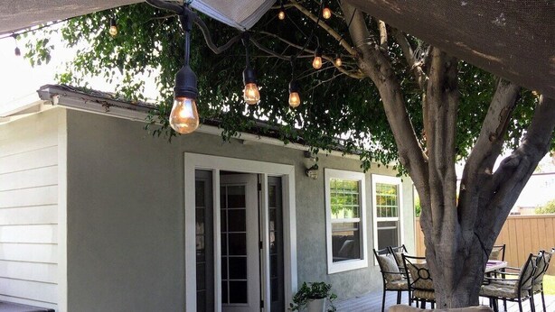 A covered outdoor seating area with a cushioned sofa, a dining table, and hanging light fixtures. - Charming Mid-Century Craftsman ~ Hot Tub/Serene Yd (San Diego)