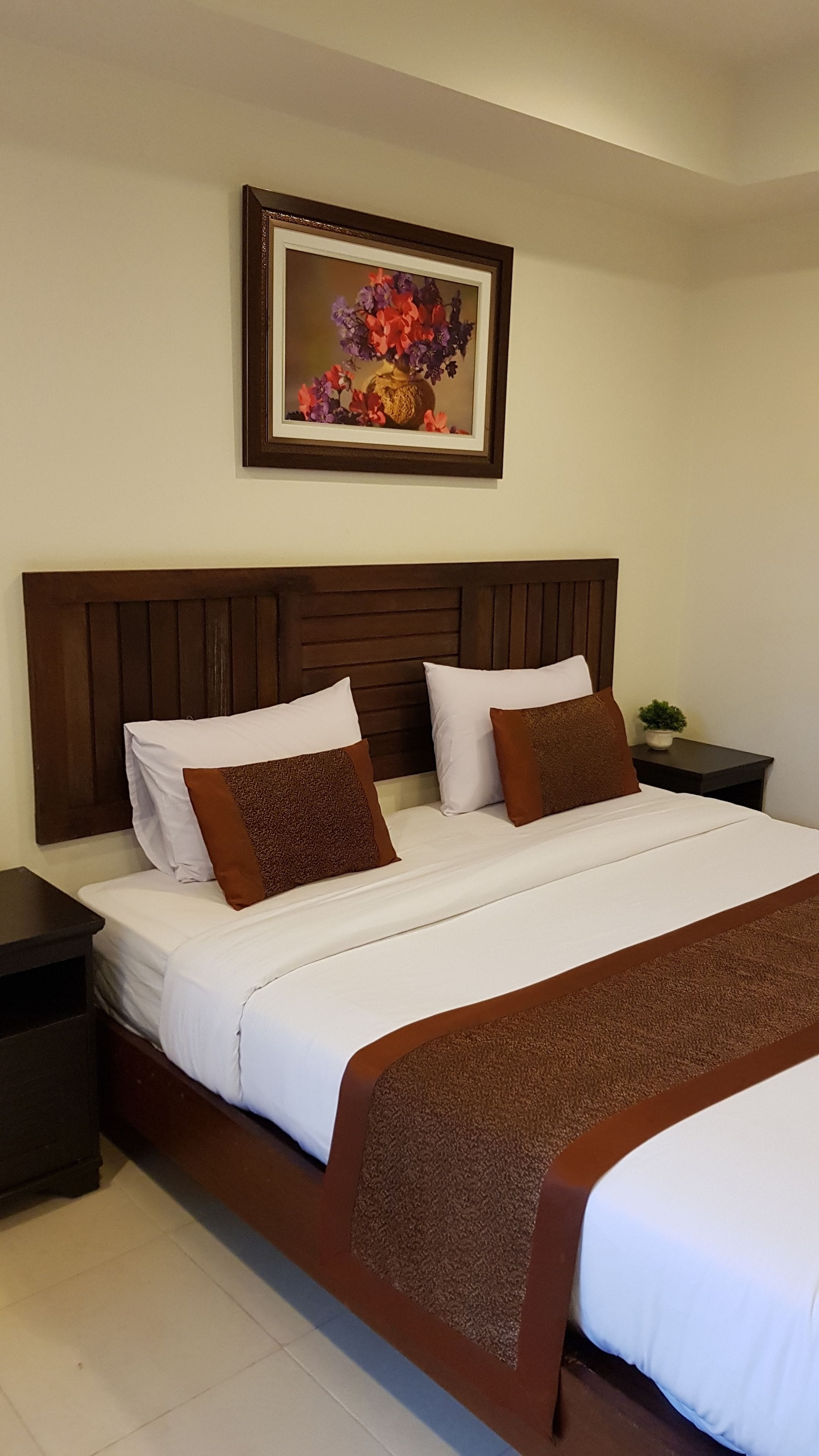 Una cama doble con ropa de cama blanca y marrón, un cabecero de madera y una mesita de noche con una planta.