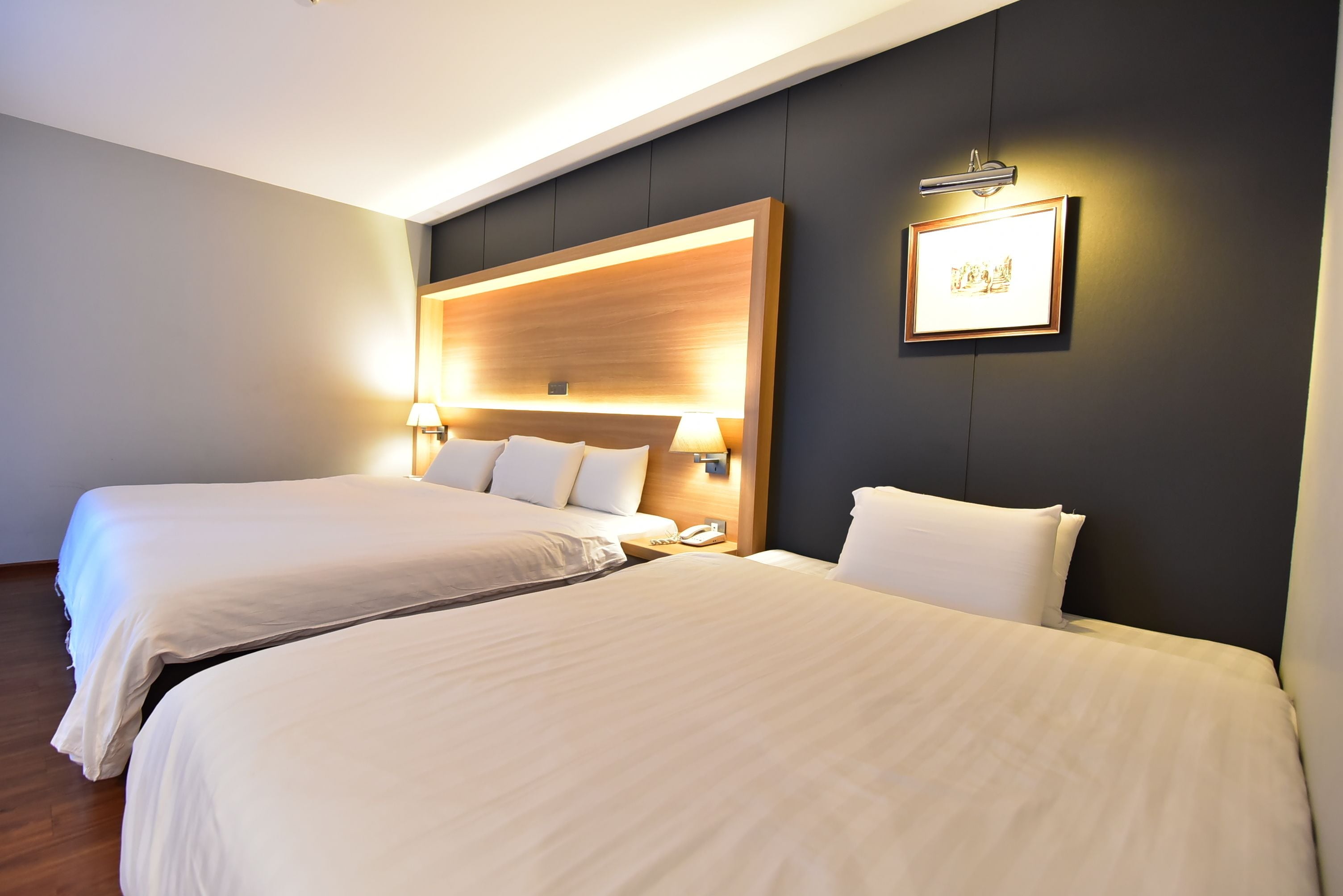 A hotel room with two beds, a wooden headboard, bedside lamps, a framed picture, and a phone on the nightstand.