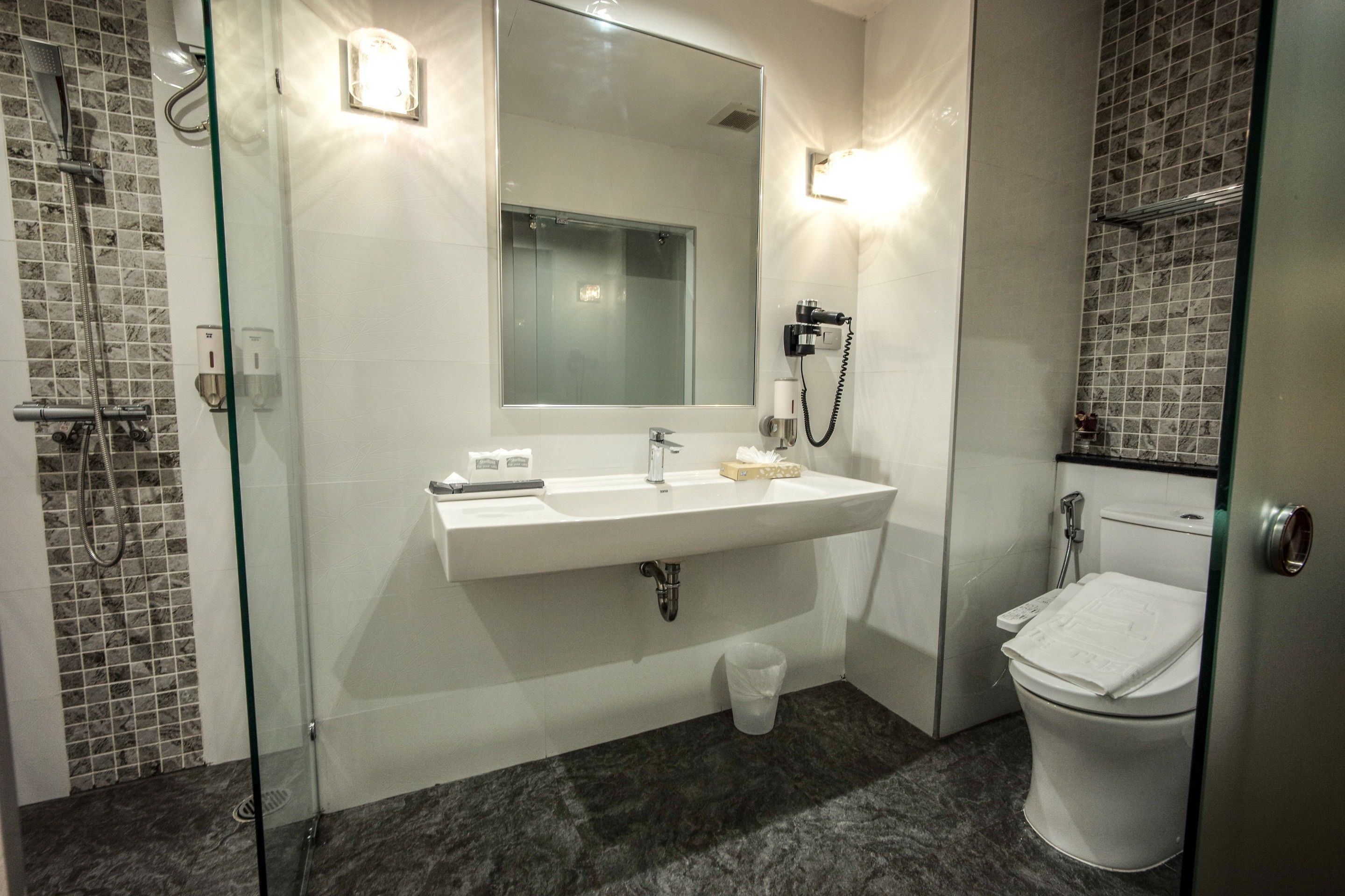 A modern bathroom with a glass shower enclosure, a white sink with a mirror above it, and a toilet with a paper towel holder.