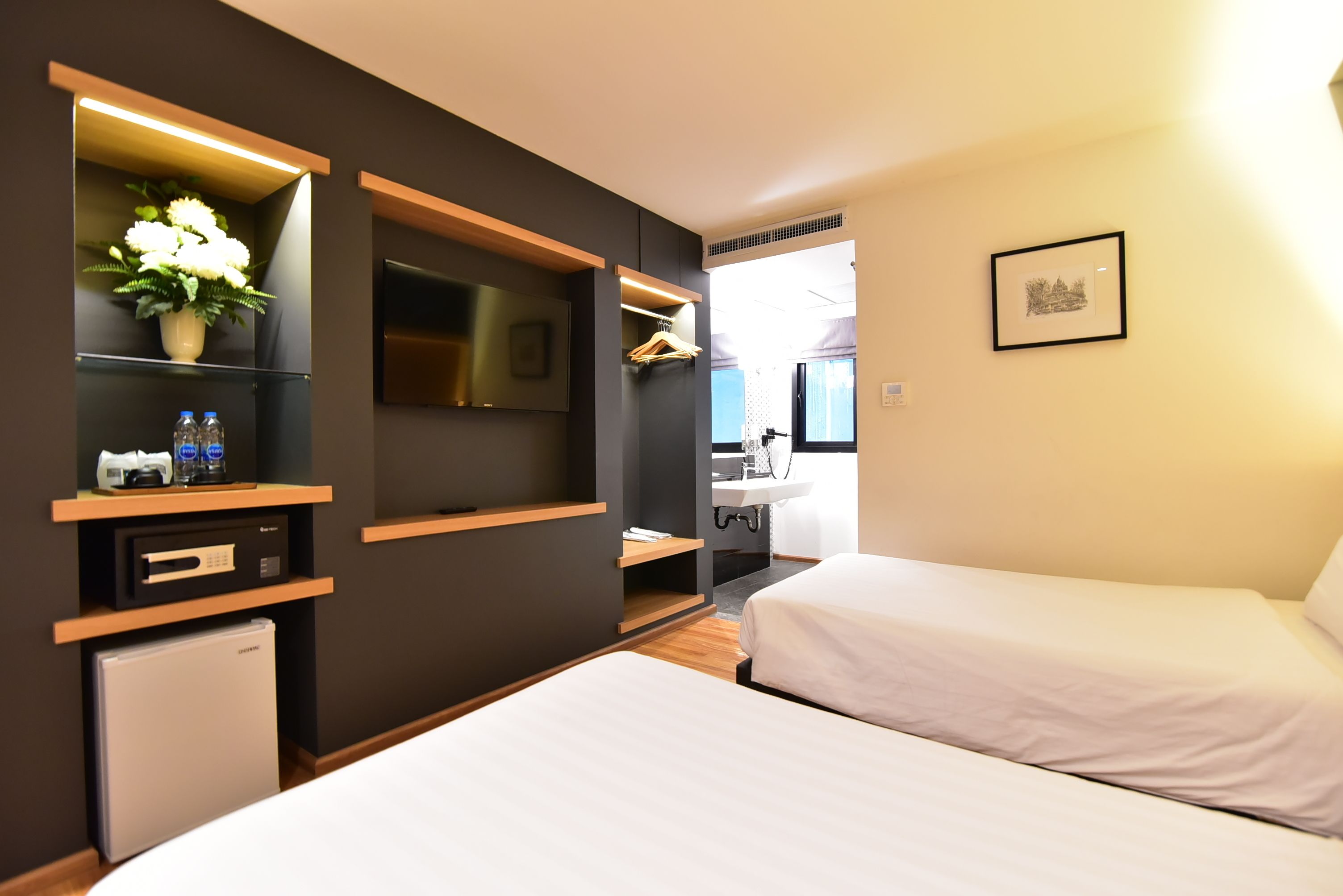 A modern hotel room with two beds, a small refrigerator, a TV, a plant, and a bathroom visible in the background.