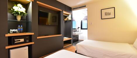 A modern hotel room with two beds, a small refrigerator, a TV, a plant, and a bathroom visible in the background.