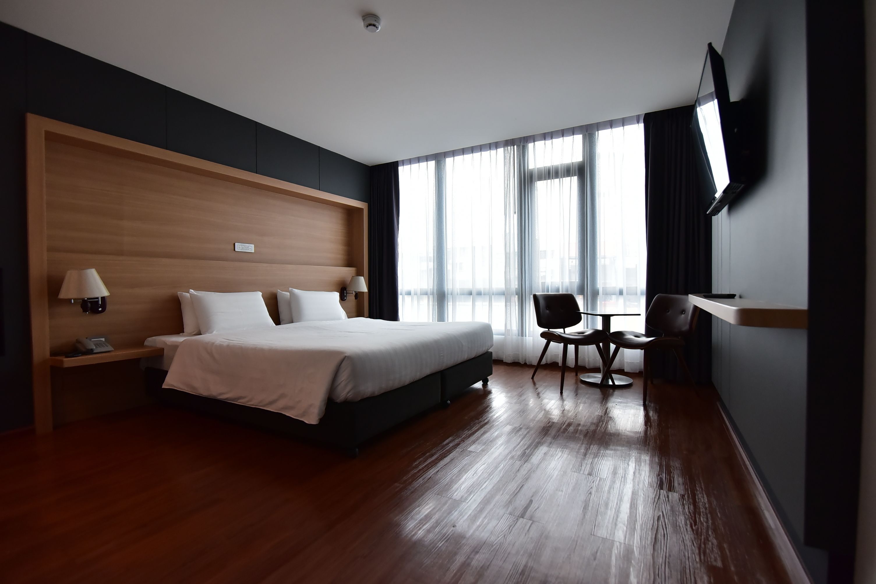A modern hotel room with a large bed, a desk with a chair, a TV mounted on the wall, and large windows with curtains.