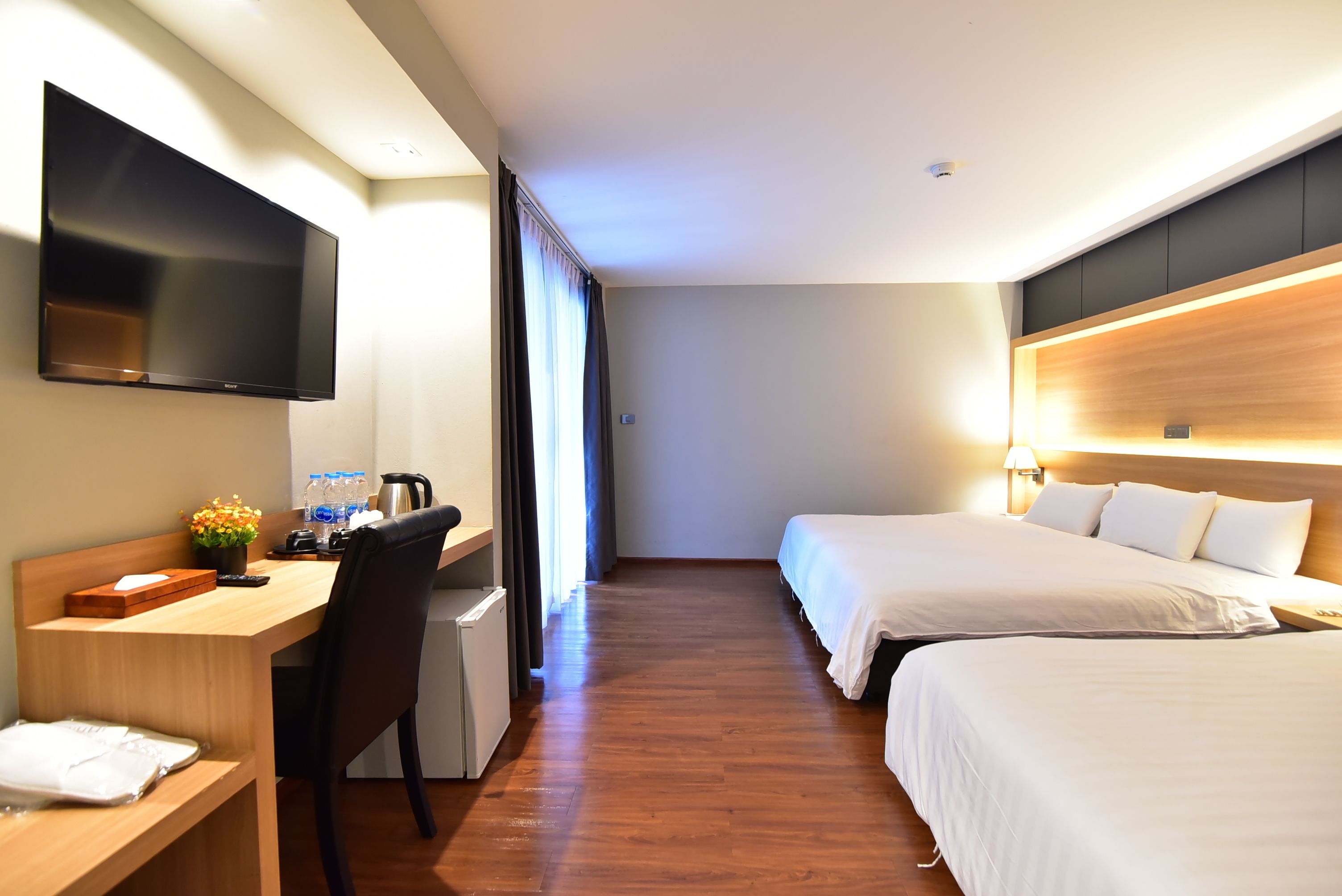 A hotel room with a wooden desk, a flat-screen TV, two beds, and a large window with curtains.