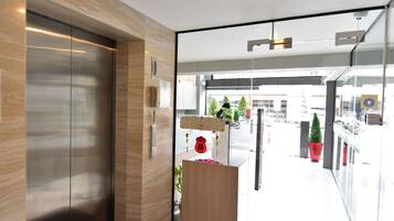 A modern elevator with a wooden finish and a glass door leading to a lobby area with a reception desk, potted plants, and a view of the street outside.