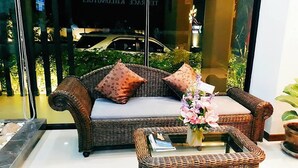 A wicker sofa with cushions, a small table with a vase of flowers, and a glass window reflecting a car outside. - The Terrace Khlongtoei (Hat Yai)