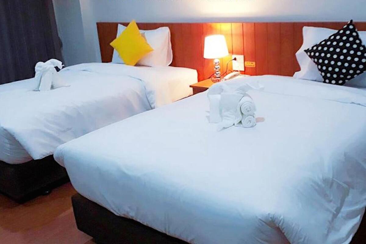 A hotel room with two single beds, each with white linens and pillows, a wooden headboard, and a bedside table with a lamp.