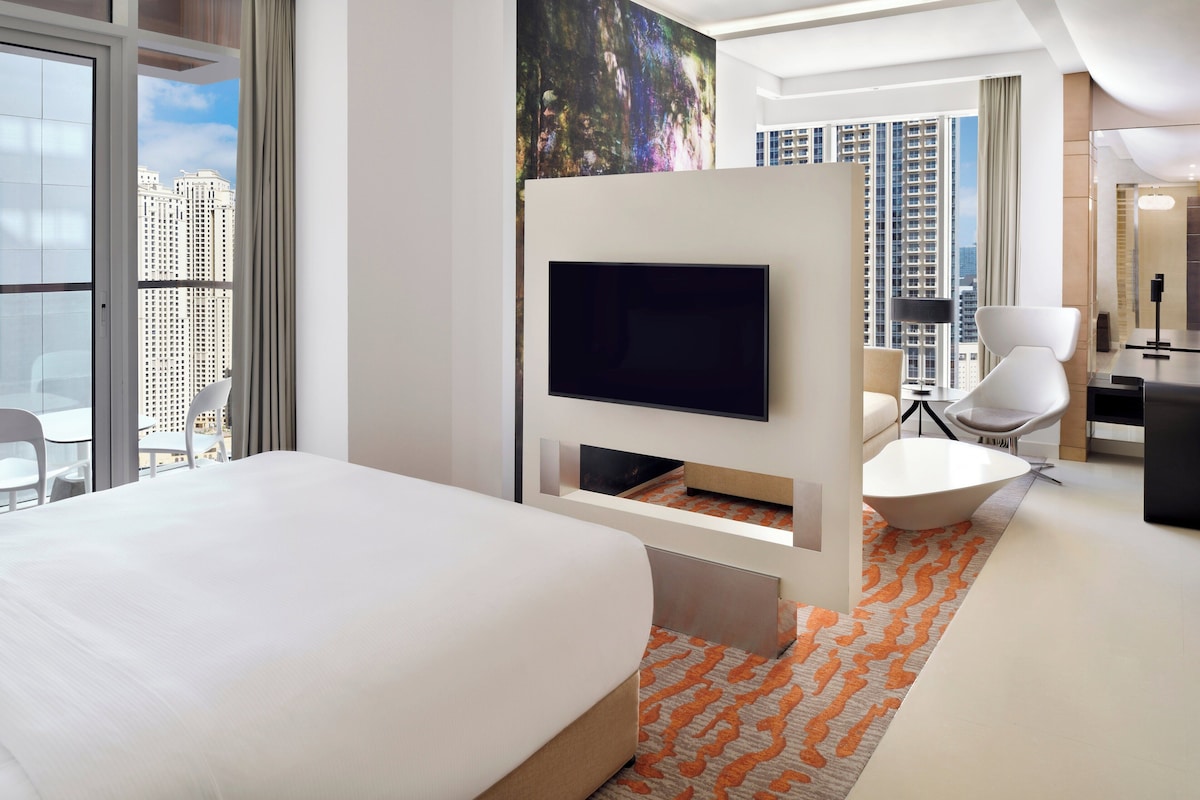 A modern hotel room with a large bed, a flat-screen TV, a white armchair, and a view of the cityscape.