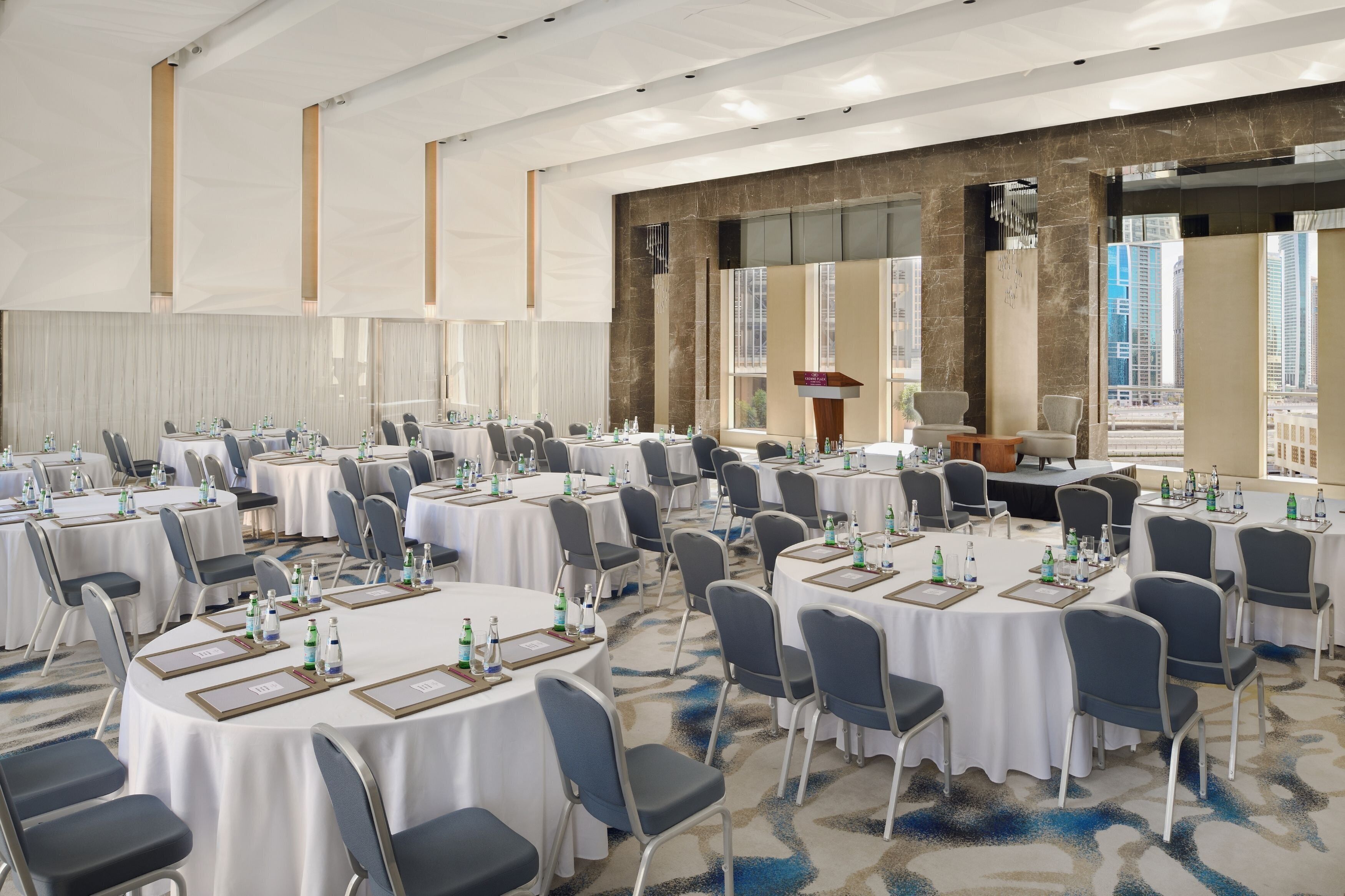 Ein Konferenzraum mit mehreren runden Tischen, an denen Namensschilder, Wasserflaschen und Notizblöcke bereitstehen. Im Raum befindet sich ein großes Fenster mit Blick auf Gebäude, und im Hintergrund ist ein Rednerpult zu sehen.