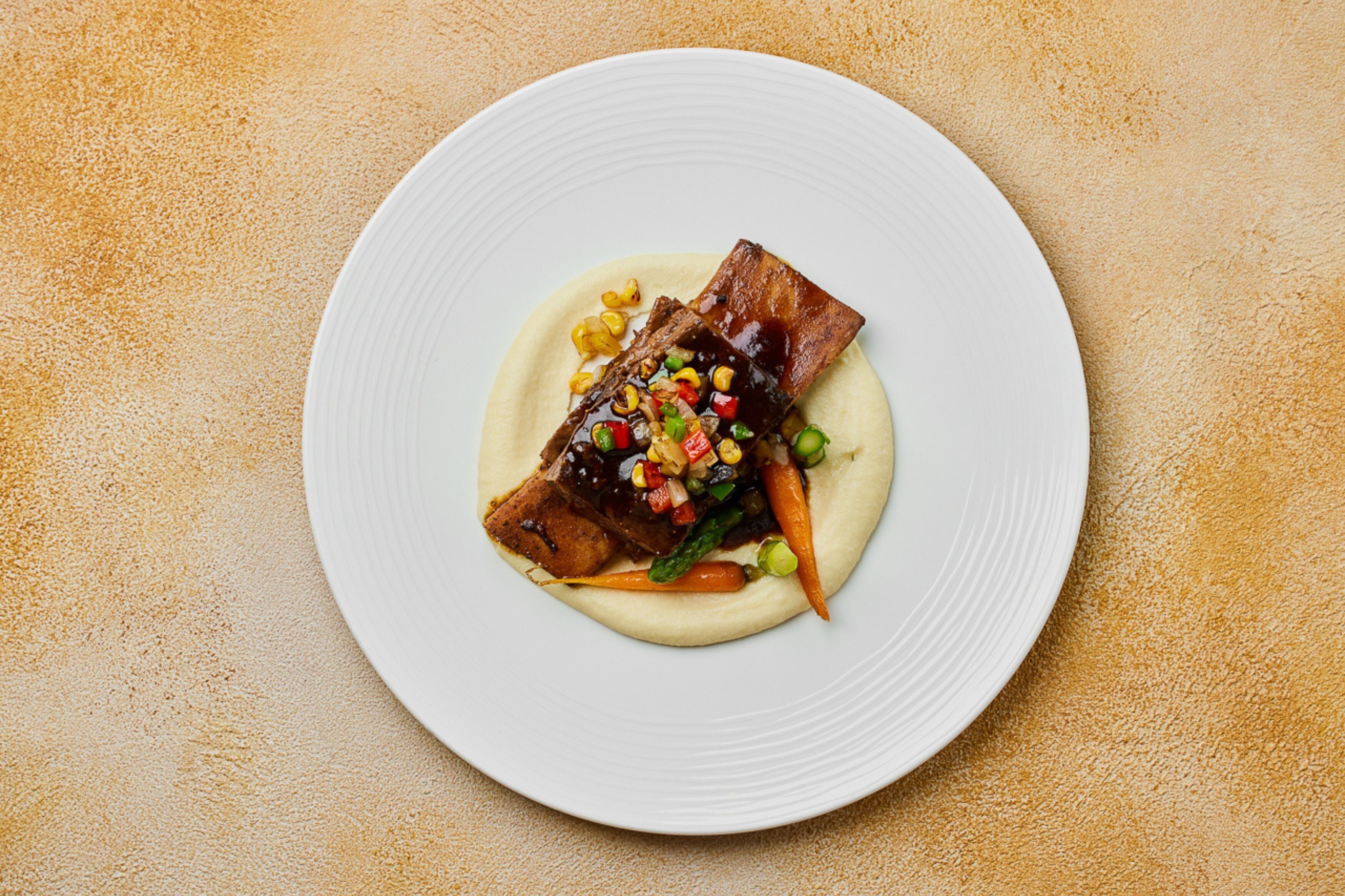 Un plato blanco con carne, verduras y una salsa cremosa sobre una superficie naranja texturizada.