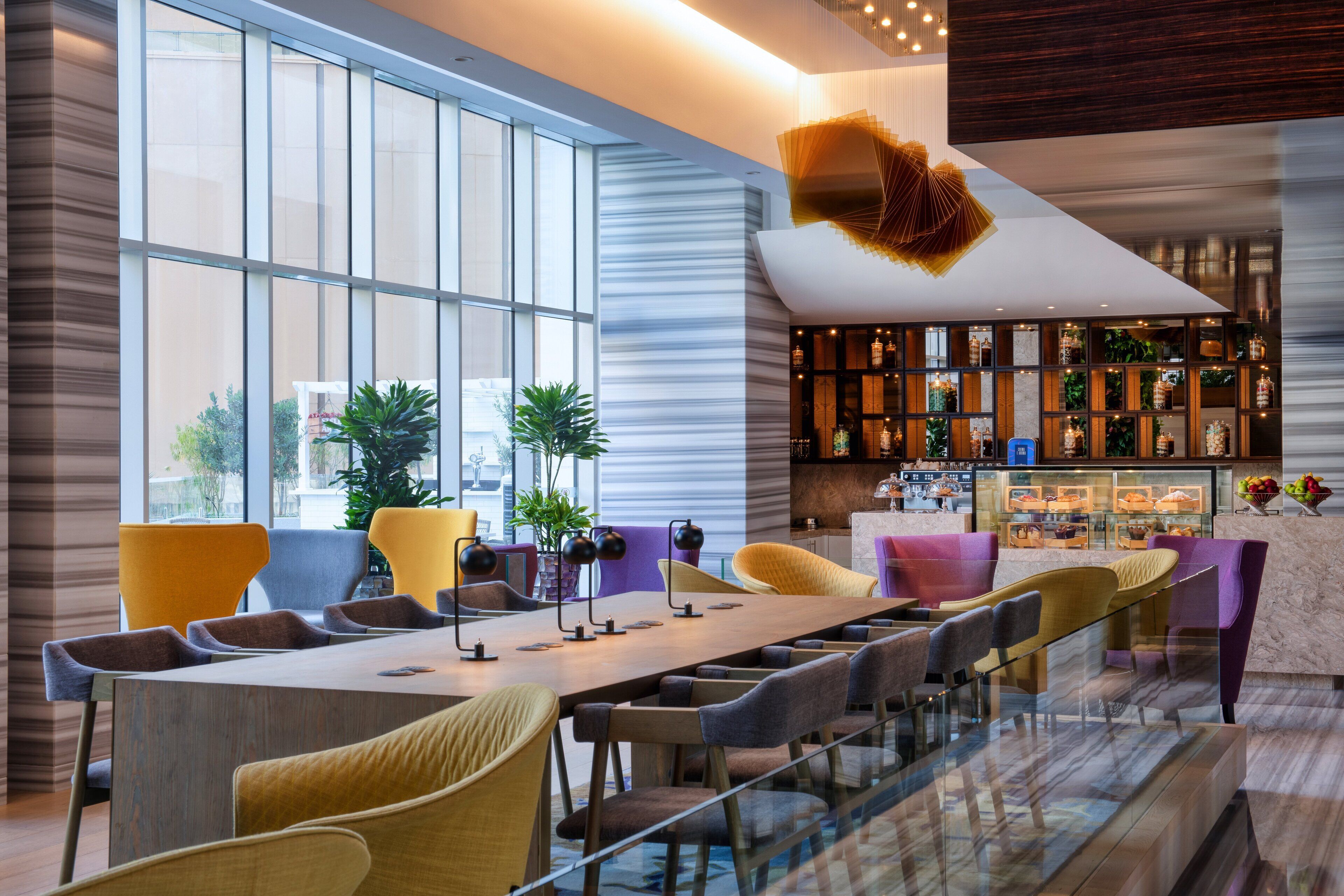 A modern dining area with a long table, colorful chairs, and a glass partition. There are potted plants and a bar area with shelves in the background.